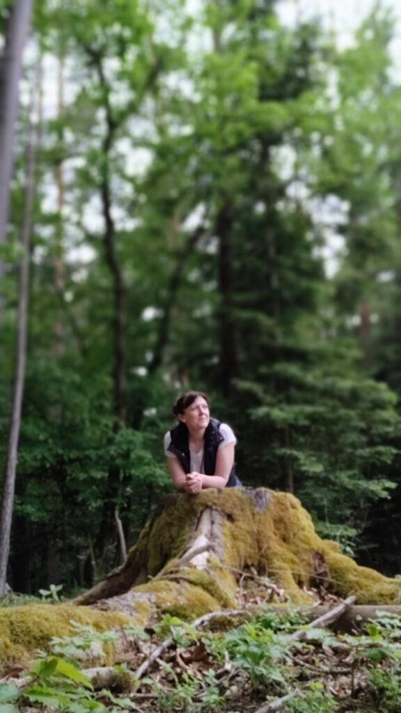 TEM Expertin Astrid Marion im Wald, die Natur beobachtend an einen Baumstumpf gelehnt